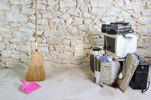 The image depicts a small collection of discarded household and electronic waste items placed on a concrete floor against a rustic stone wall. On the left, a traditional straw broom with a wooden handle leans upright, with a bright pink plastic dustpan lying flat on the ground nearby. To the right, there is a stack of unwanted electronic appliances, including an old CRT computer monitor, a tower-style desktop computer, a beige landline telephone, and a number of tangled cables and wires. A standard two-tone off-white and grey desktop fan is positioned next to the electronics. The scene suggests a cluttered area where rubbish, likely destined for removal, is temporarily stored, aligning with services offered by Commercial Waste Abbey Wood in the local area near Abbey Wood, London. The background features a textured stone wall, and the overall lighting appears natural, highlighting the variety of objects and materials in the collection meant for waste clearance and disposal.