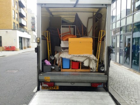 The image shows the open rear of a commercial waste collection vehicle parked on a city street, with the loading area filled with various household items and rubbish, including a yellow storage box, a foldable trolley with orange handles, a black bag, and a blue container. A grey fluffy rug partially covers some items on the lower level. The backdrop features a modern glass-fronted building on the right side and residential or commercial buildings on the left, with a pavement running alongside. The vehicle's licence plate, 'KUB1LFV,' is visible at the bottom. This scene, associated with rubbish removal services, reflects an urban environment in or near Abbey Wood, and the contents are representative of domestic or light commercial waste collection typical for companies like Commercial Waste Abbey Wood.