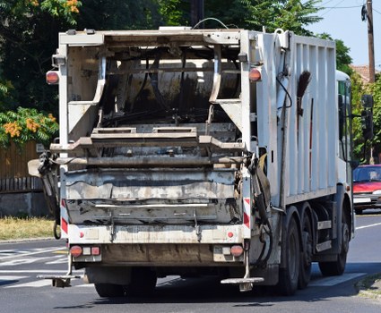 A white commercial rubbish collection truck with a large, rectangular rear compactor is depicted driving along a suburban street in daylight. The rear section of the truck shows accumulated debris and waste materials, with visible layers of dirt, grime, and some faded black and grey stains on the metal surfaces. The truck’s structure includes hydraulic arms and mechanisms for loading and compacting waste, with safety reflectors and red-and-white striped warning markings along the bottom edges. In the background, there are green trees, a residential wooden fence, and a few parked vehicles, indicating a typical urban or suburban environment near Abbey Wood. The roadway is paved, and the scene is well-lit by natural sunlight, highlighting the utilitarian appearance of the waste collection vehicle as part of municipal rubbish removal services. Commercial Waste Abbey Wood provides local rubbish collection, and this truck exemplifies its fleet used for municipal and commercial waste management in the area.