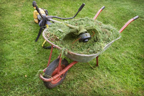 A red, manually operated wheelbarrow filled with a large amount of cut grass and garden debris, including grass clippings and small plant remnants, situated on a well-maintained grassy area. The wheelbarrow's metal tray appears weathered and partially covered by the green clippings, with the handles extending upwards and outwards, one of which shows signs of slight rust. Behind the wheelbarrow, there is a yellow and black petrol-powered grass trimmer or brush cutter resting on the grass, with its handle and control cables visible. The scene is outdoors, likely in a garden or backyard, with natural daylight illuminating the area evenly, typical of a residential or small commercial setting in the Abbey Wood area, illustrating typical garden waste collection or disposal activities that a waste management company like Commercial Waste Abbey Wood might handle for local clients in the postcode area.