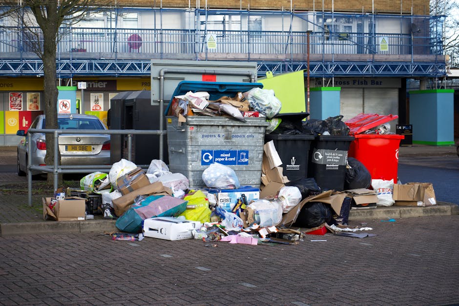 A middle-aged man with dark hair, wearing a black T-shirt with white text and dark trousers, is seen disposing of a white plastic bag filled with rubbish into a tall, cylindrical stainless steel waste