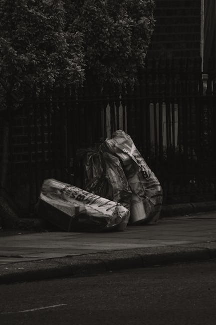 Two large black garbage bags, made of plastic with a shiny, crinkled texture, are placed on the edge of a paved street near the curb. The bags appear to be filled with waste materials and are tied at 