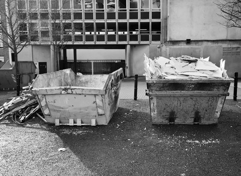 Two large industrial-style skips positioned on a paved area outside a building in Abbey Wood, with one skip filled with what appears to be crumpled and torn paper or cardboard, while the other contains mixed waste materials. The skip on the left is made of metal with a white finish, showing signs of rust and wear, and is partially filled with debris, including possibly broken wooden pallets leaning against it. The skip on the right is darker, possibly black or dark gray, and is filled with irregularly stacked paper and cardboard, some overflowing onto the ground. In the background, a modern concrete building with a grid-style façade and small windows is visible, along with a metal gate beneath an overhang. The area appears to be a commercial or industrial site, with sparse surroundings and no visible vegetation, consistent with rubbish removal operations in Abbey Wood, southeast London. Commercial Waste Abbey Wood manages waste collection and disposal in this urban setting, serving local businesses with reliable rubbish clearance services.