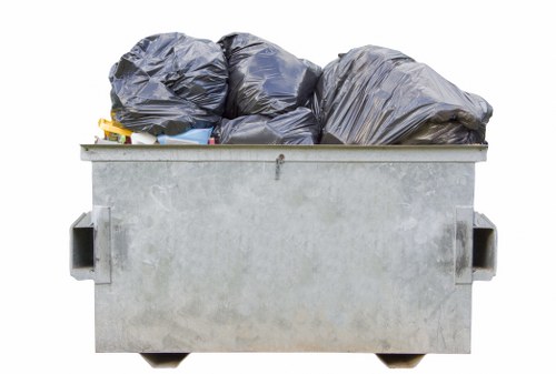 A large, industrial-sized metal dumpster positioned outdoors on a paved surface, filled with multiple tightly packed black rubbish bags made of plastic, exhibiting a slightly crinkled texture. The bags are stacked on top of each other, some leaning slightly to the sides, covering the top of the container completely. The dumpster has a plain, silver-grey finish with reinforced handles on each side and small wheels visible at the bottom. In the background, there are no other objects or structures visible, suggesting a clear, open environment typical of commercial waste disposal grounds in Abbey Wood. The overall scene is well-lit with natural daylight, highlighting the metallic surface of the container and the glossy surface of the bags. This image visually represents rubbish collection and waste management services provided by Commercial Waste Abbey Wood for commercial or industrial clients in the local area, fitting with a webpage about effective commercial waste solutions in Abbey Wood.