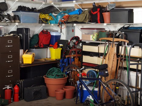 A cluttered garage or storage room filled with various household items and rubbish. In the foreground, there are multiple plant pots made of terracotta and plastic, some stacked and others placed on the floor, with a garden hose coiled up nearby. A blue bicycle with a black seat and handlebars is positioned against a shelving unit, partially obscuring it. The shelves above and behind feature plastic storage boxes, tools, and miscellaneous items, including a red and black bag, a grey container, and a yellow plastic box. On the left, a black filing cabinet is visible, while the right side contains a wooden step ladder, a black stereo speaker, and a small black radio or amplifier. The background shows metal wire shelves holding more containers, cords, and household equipment, with a bright light illuminating the space. The overall scene depicts a typical domestic storage area used for rubbish accumulation awaiting disposal, consistent with services offered by Commercial Waste Abbey Wood in the local postcode area.
